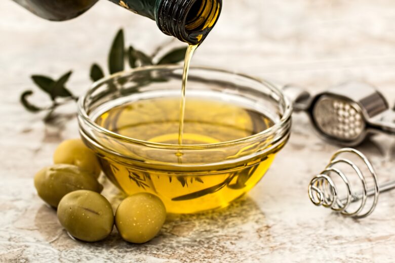 Medbest extra virgin olive oil being poured into a glass bowl with green olives and kitchen utensils on a marble surface